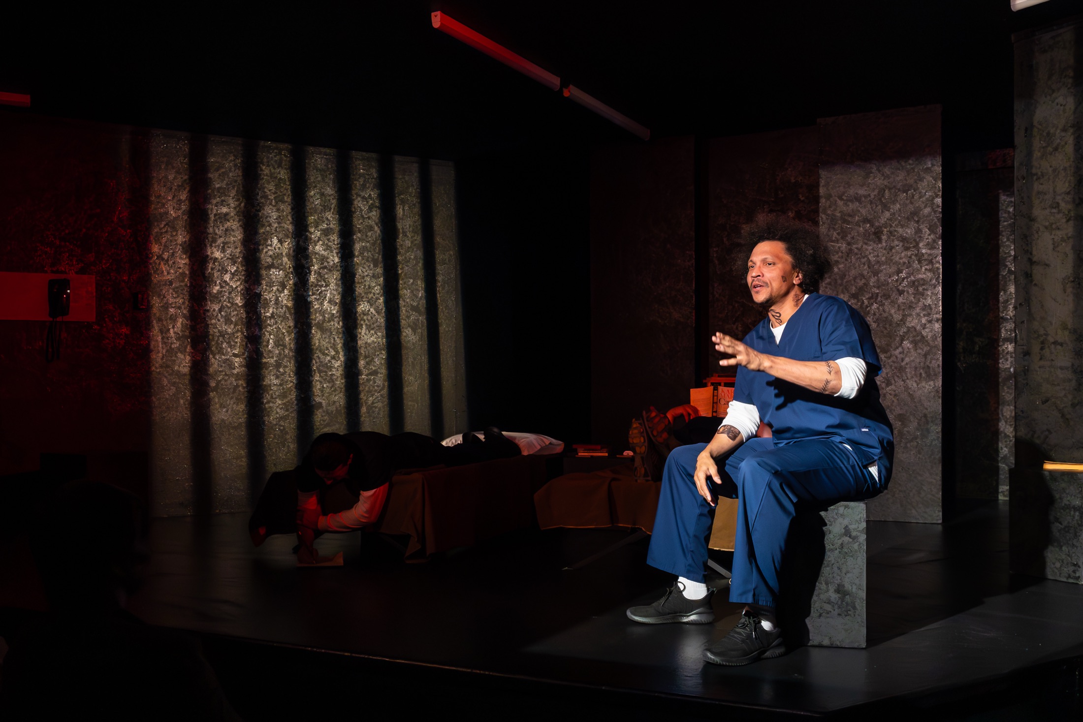 A performer in blue scrubs speaks while seated on the prison-stage set